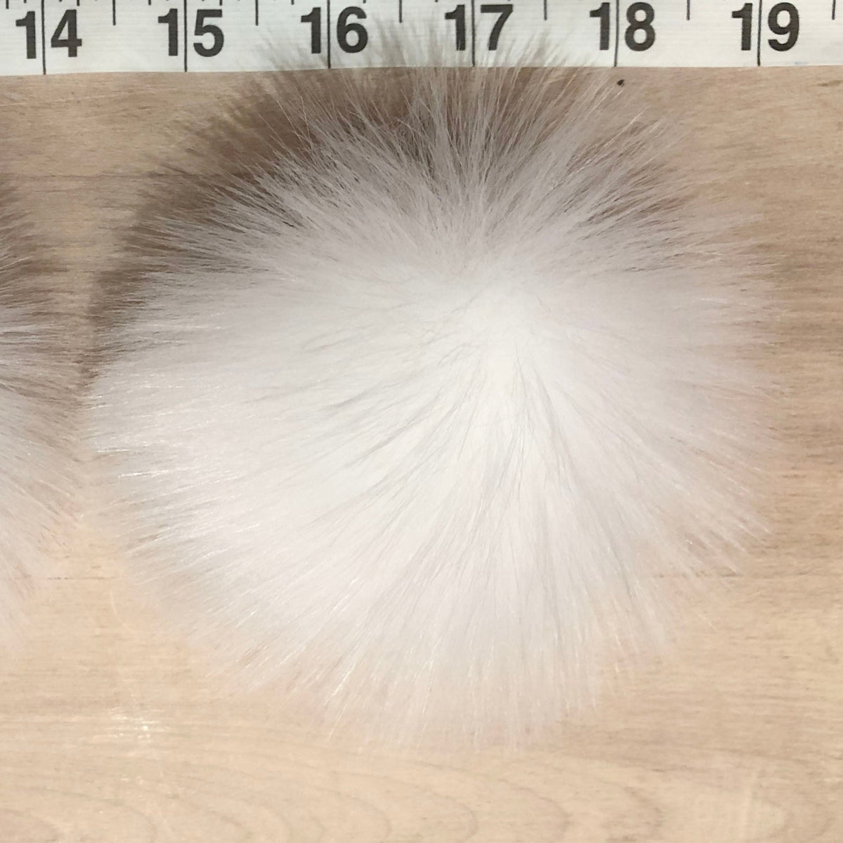 Two fluffy white faux fur pom-poms placed on a light wood surface with a measuring tape visible at the top for scale.