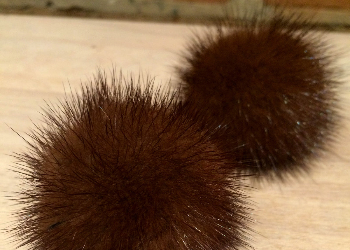 Two dark brown furry pom-poms on a light wood surface with a brick wall in the background.