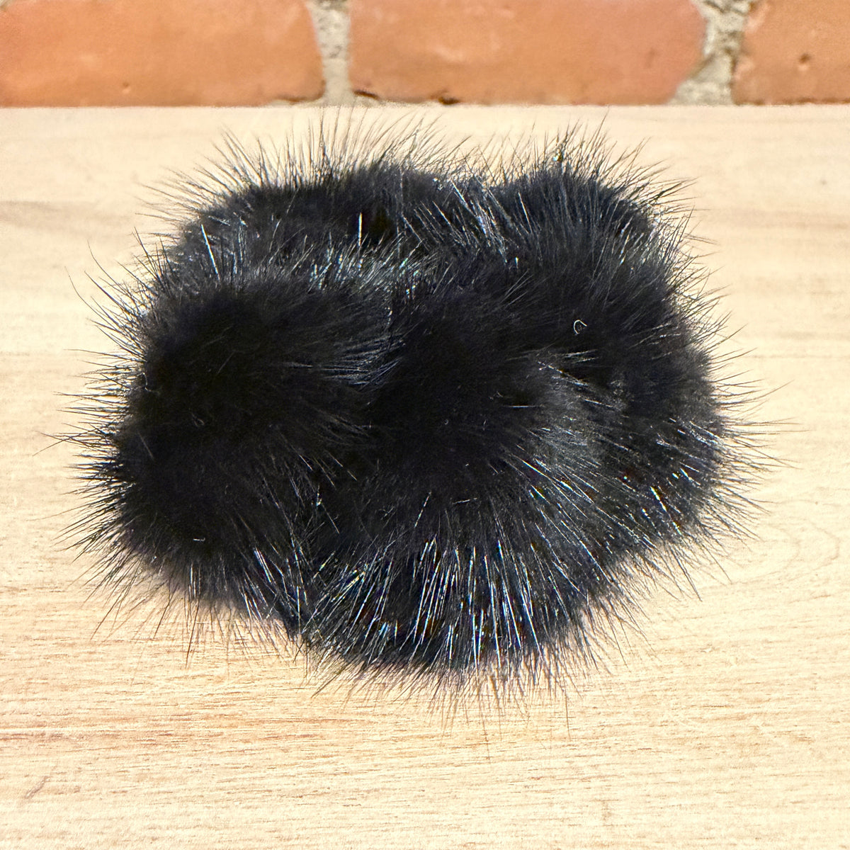 A black fluffy mink fur hair accessory, likely a pom pom scrunchie, resting on a light wood surface with a brick wall in the background.