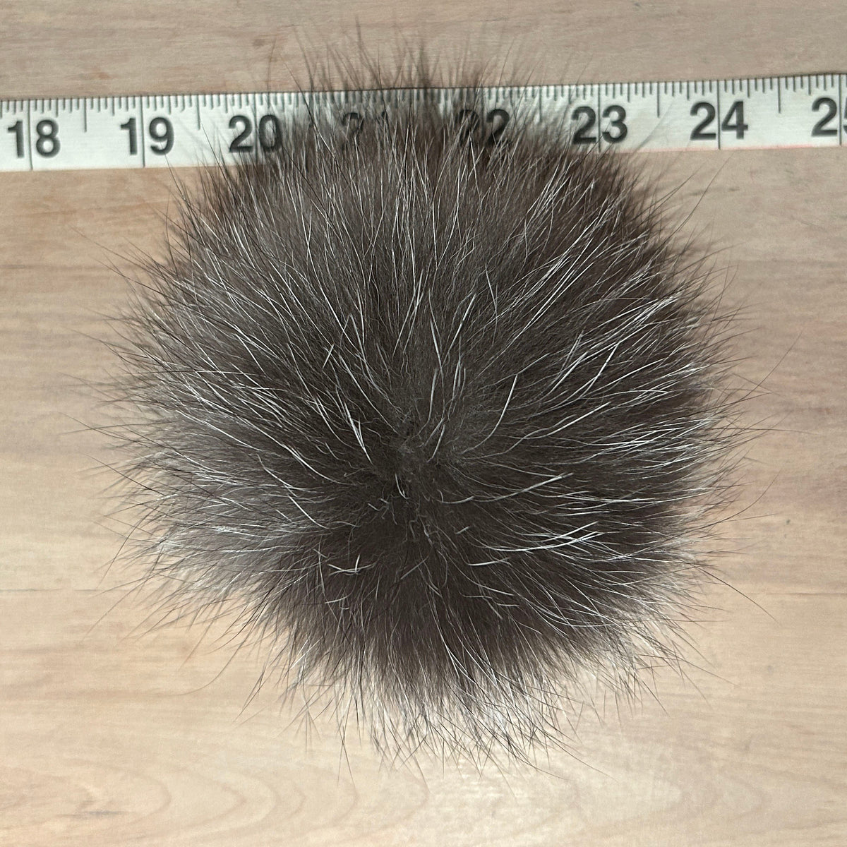A round, fluffy fox fur pom-pom in a dark brown and gray colorway, shown on a light wood surface with a measuring tape ruler at the top to indicate size.