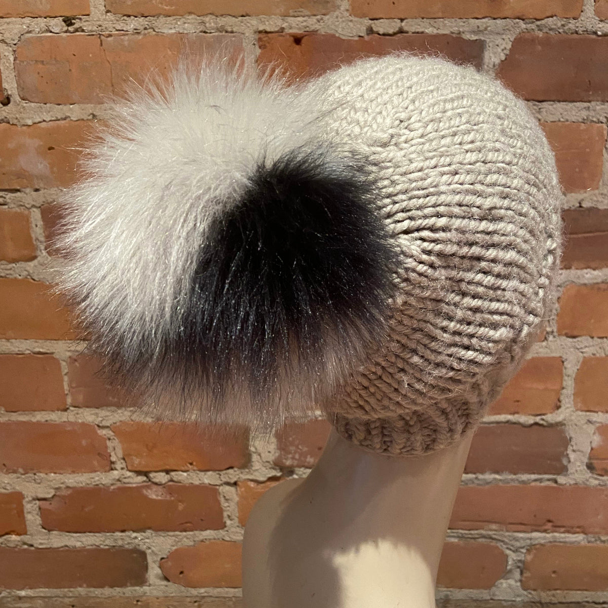A cream-colored knit beanie with a large black and white fluffy faux-fur pom-pom, displayed on a mannequin head against a red brick wall background.