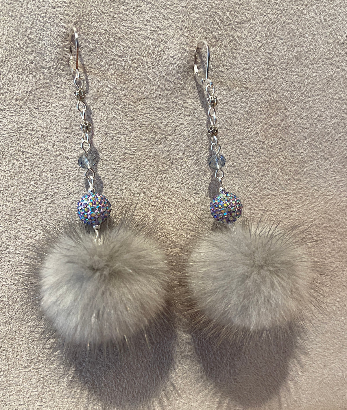 A pair of dangling earrings with silver fishhook wires, small faceted beads, iridescent blue-purple rhinestone balls, and large fluffy light gray pom-poms, displayed on a textured beige surface.