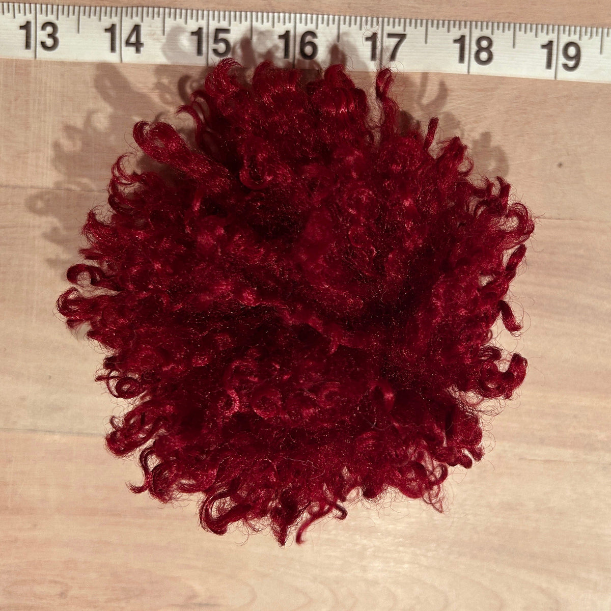 A dark red curly hat pom pom laid flat on a light wood surface with a measuring tape ruler visible along the top edge to show size.