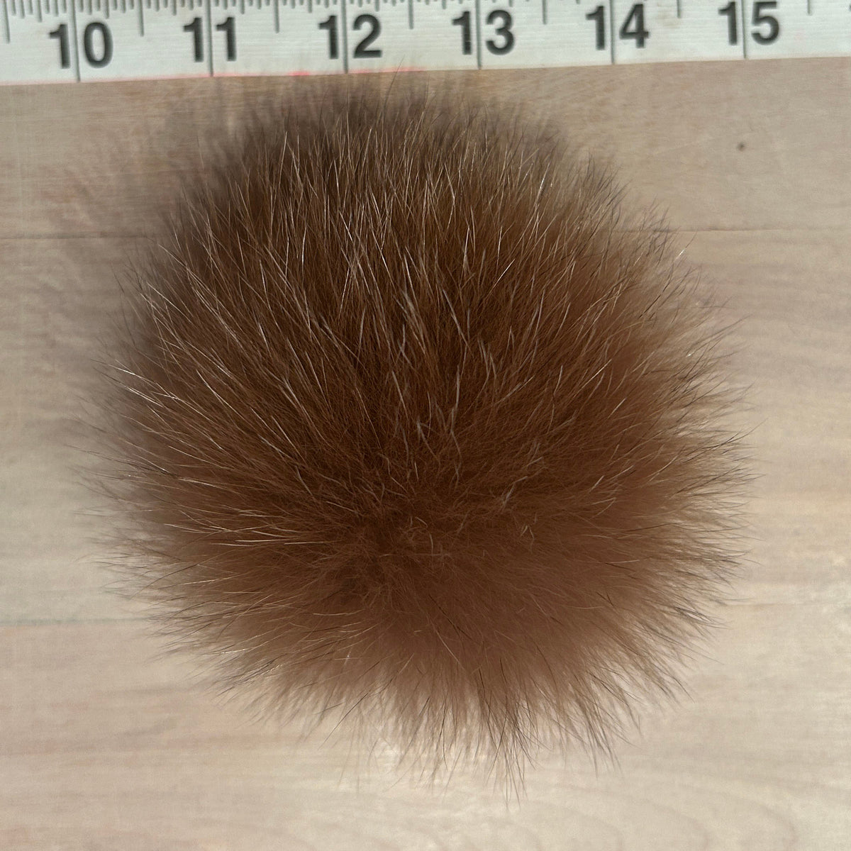 A ginger fox fur pom-pom accessory placed on a light wood surface with a measuring tape visible at the top for scale.