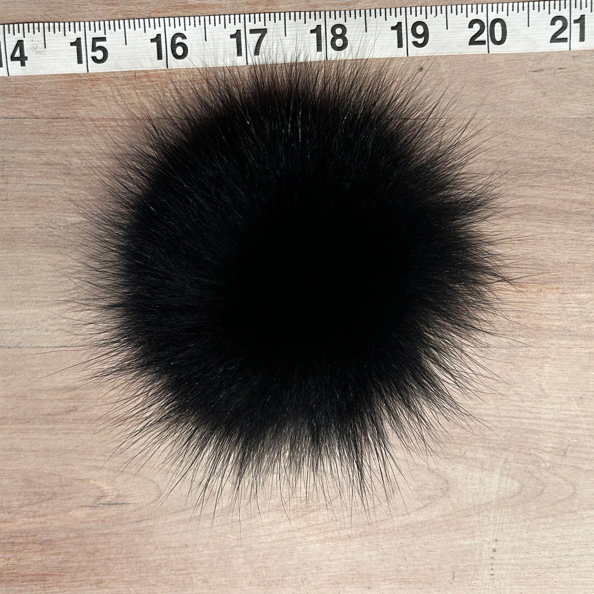 A round black fox fur pom-pom with long, spiky fibers, shown on a light wood surface with a measuring tape ruler at the top for scale.
