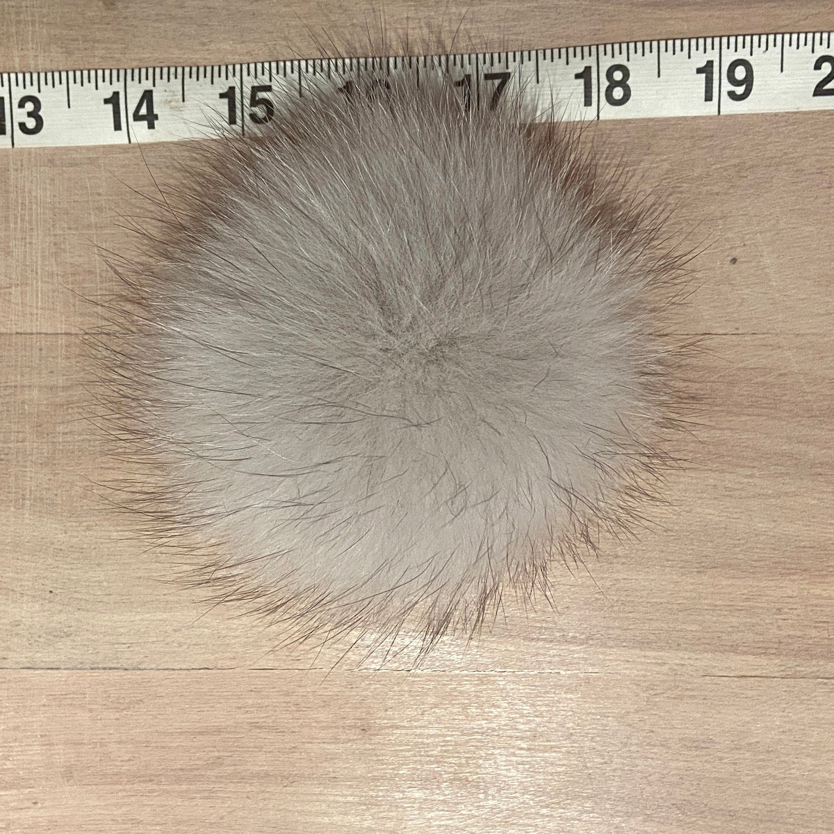 A round, fluffy fox fur pom-pom in a light gray-brown color, shown on a light wood surface with a metal ruler at the top for scale.