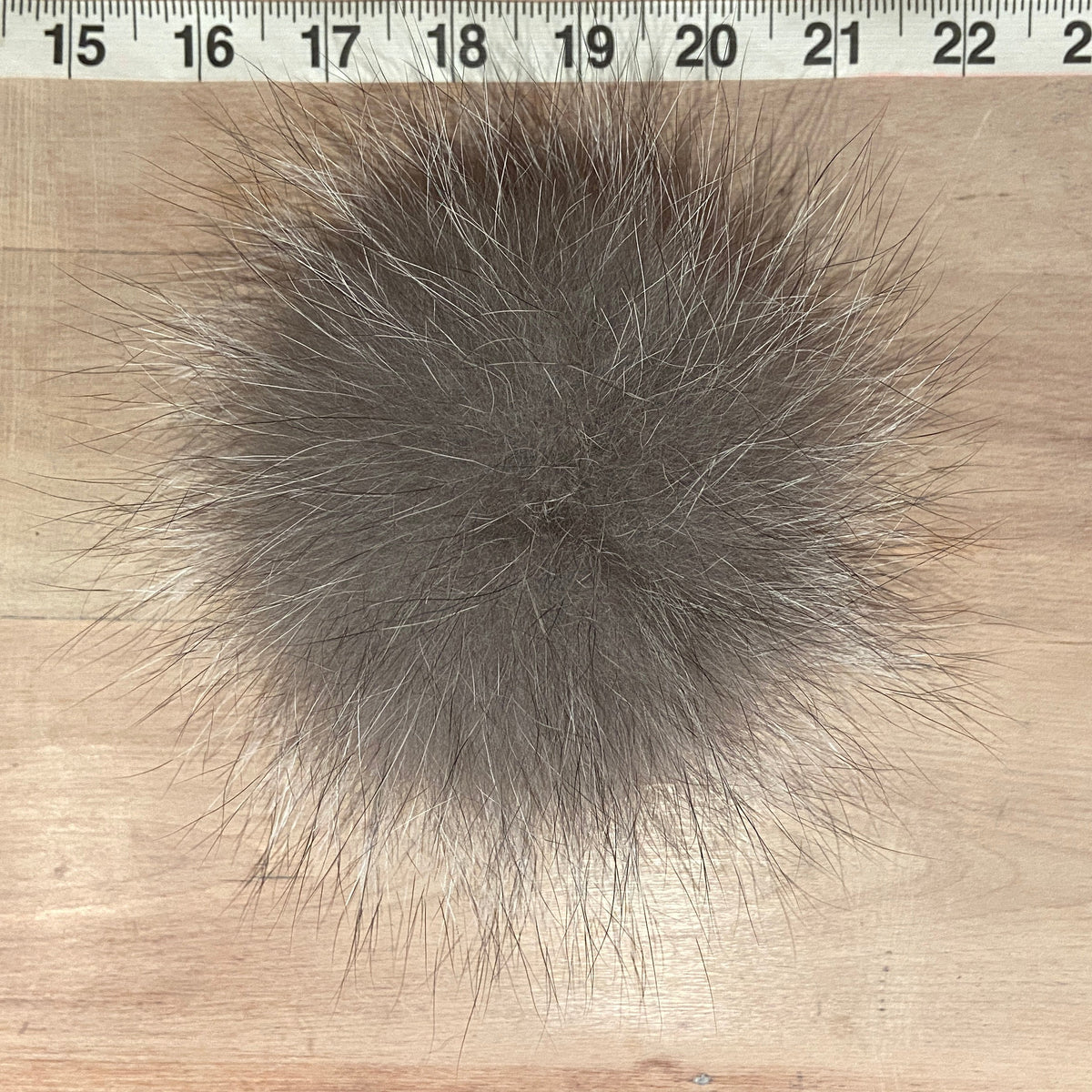 A round gray fox fur pom-pom with black and white mixed hairs, shown on a light wood background with a measuring tape ruler at the top to indicate size.