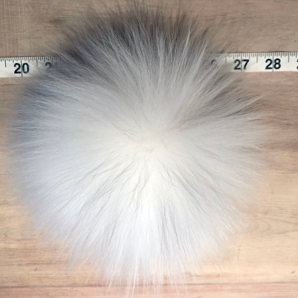 A fluffy lamb fur pom-pom with a gray and white gradient, shown on a wooden surface next to a measuring tape for scale.
