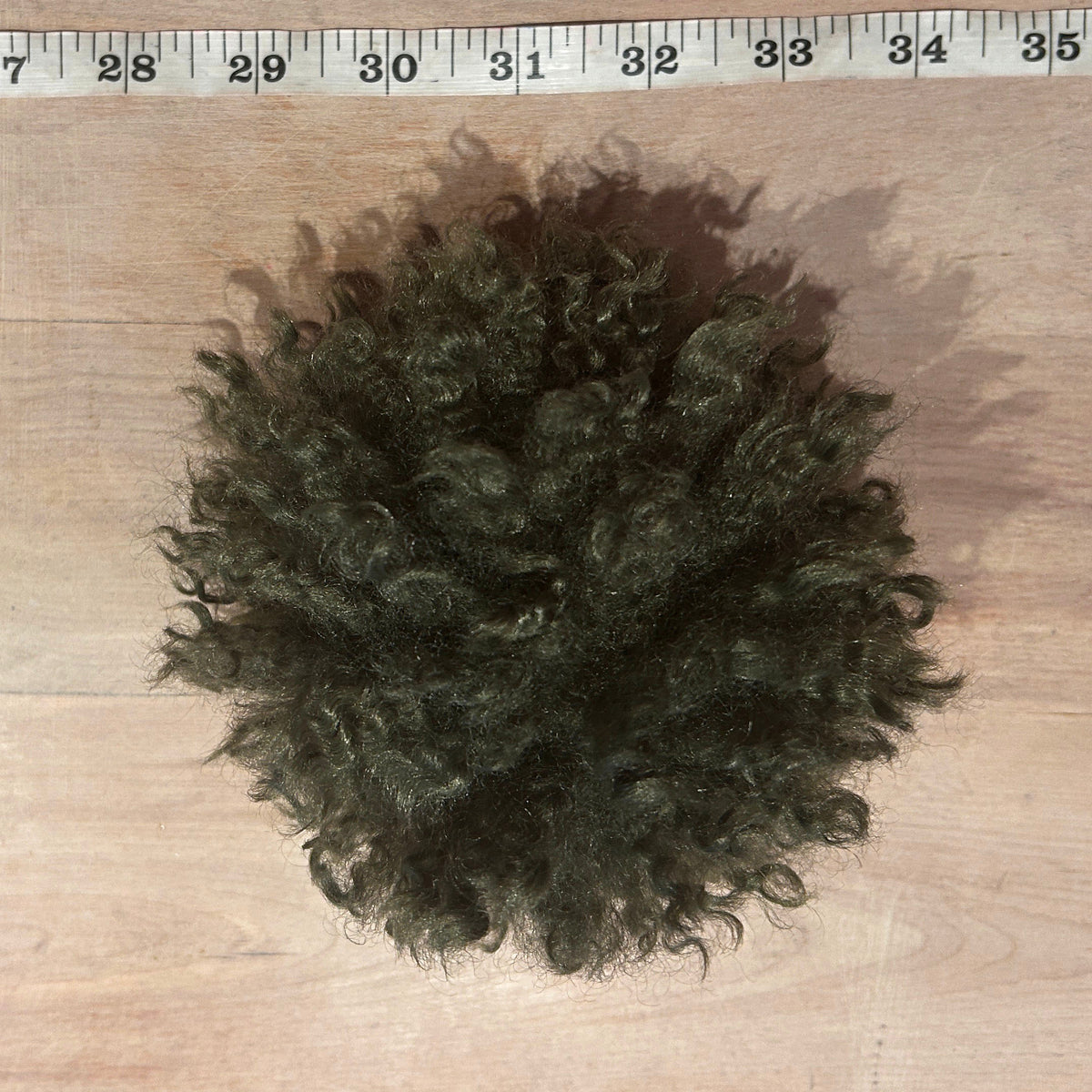 A dark olive green pom pom on a light wood surface, with a measuring tape visible along the top edge for scale.