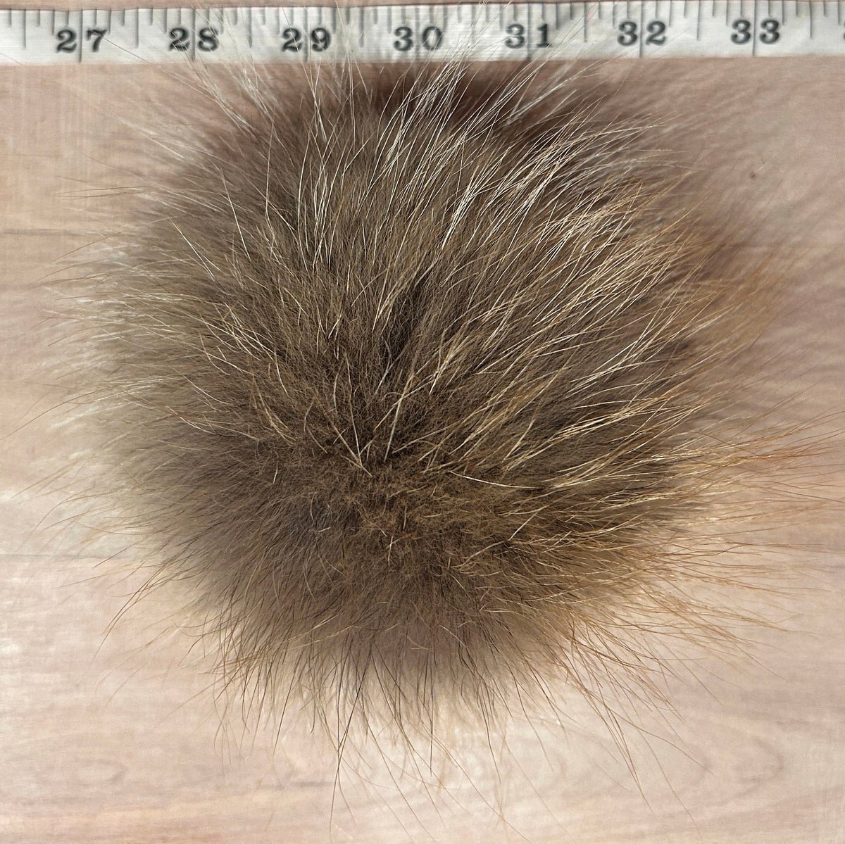 A close-up of a round, fluffy Coyote fur pom-pom in shades of brown and beige, positioned below a metal ruler to show its size against a light wood background.