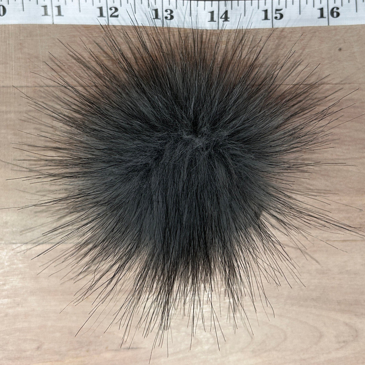 A dark gray faux fur pom-pom shown from a top-down view on a light wood surface, with a measuring tape visible at the top edge for scale.