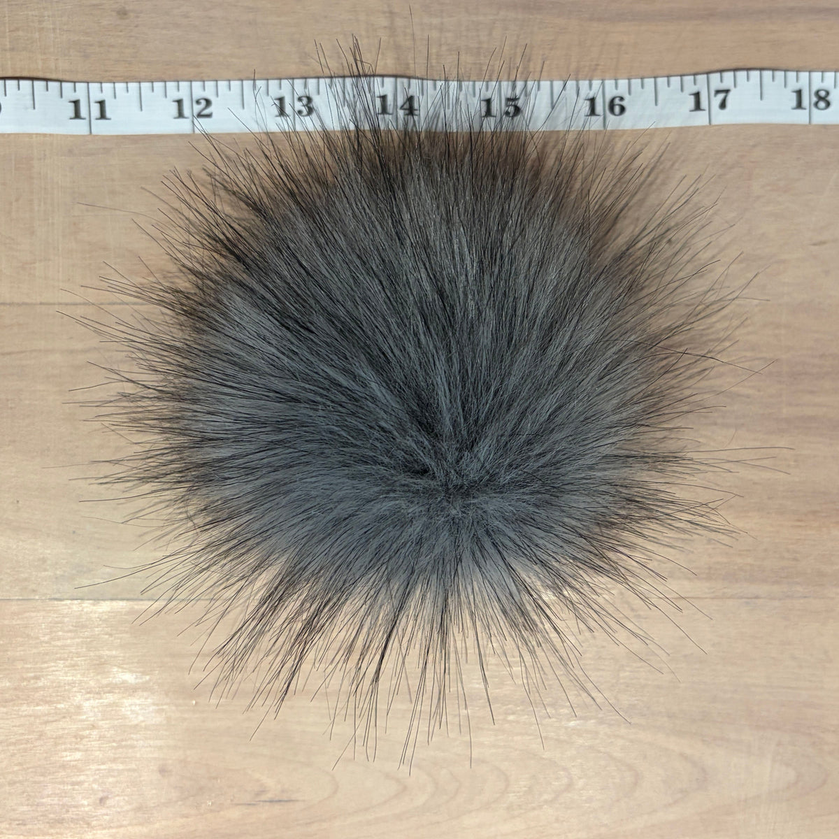 A large, fluffy dark gray faux fur pom-pom shown from a top-down view on a light wood surface, with a white measuring tape ruler positioned along the top edge for scale.