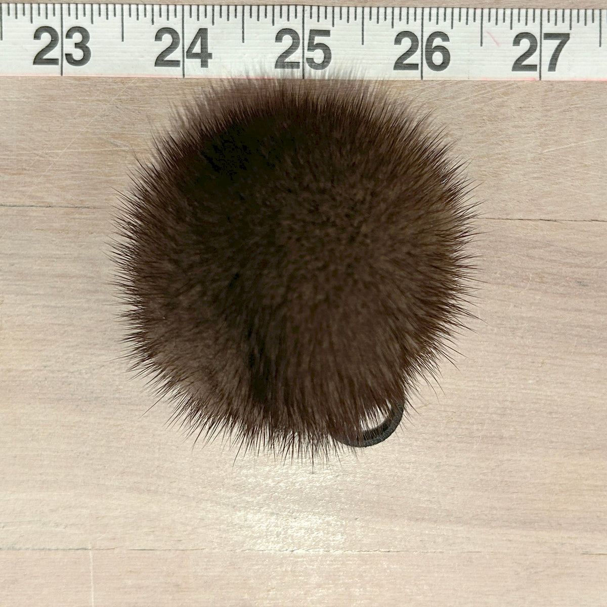 A dark brown fluffy pom-pom accessory shown from above on a light wood surface, with a measuring ruler along the top edge for scale.