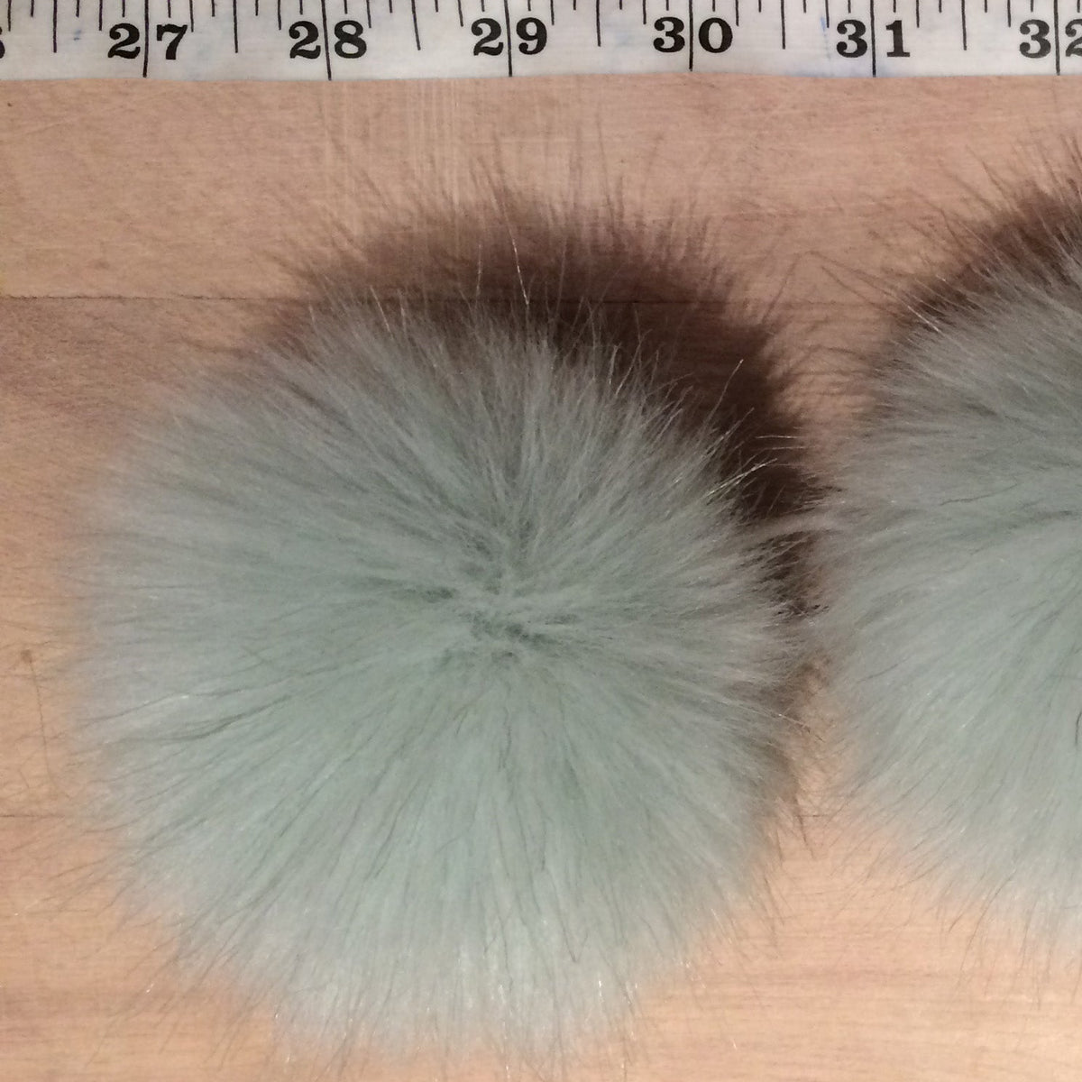 Three fluffy faux-fur pom-poms in a pale sage green color, laid out side-by-side on a light wood surface with a measuring tape visible at the top for scale.