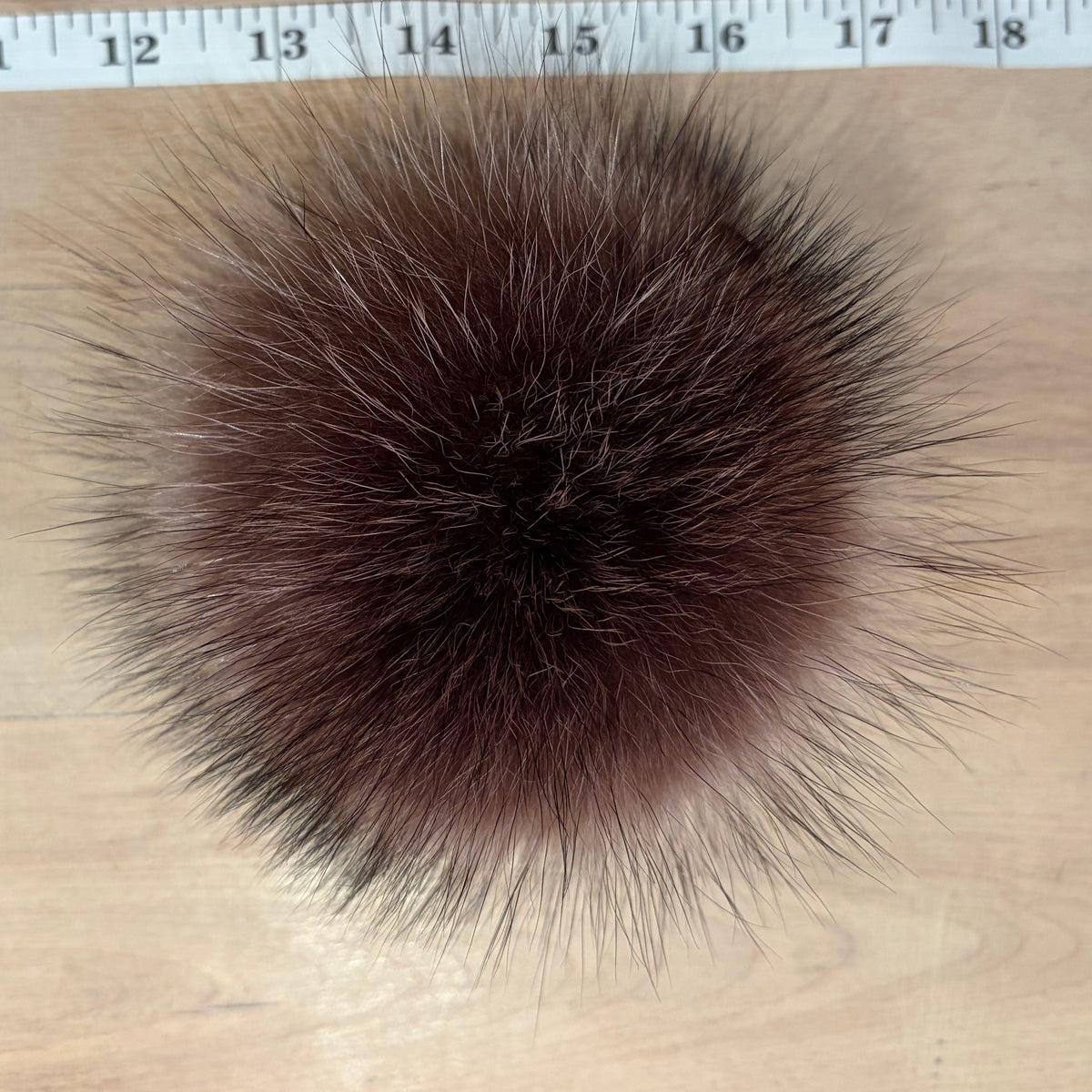 A round, fluffy raccoon fur pom-pom in a dark reddish-brown color, shown from above on a light wood surface with a measuring tape visible at the top edge.