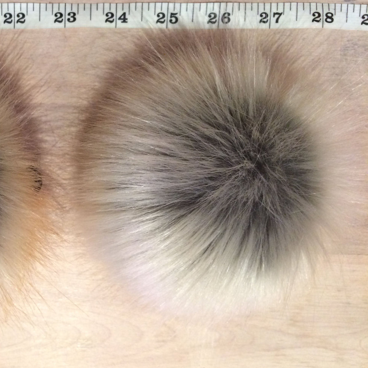 A round faux fur pom-poms with a brown, gray, and cream gradient, displayed side-by-side on a light wood background with a measuring ruler at the top.