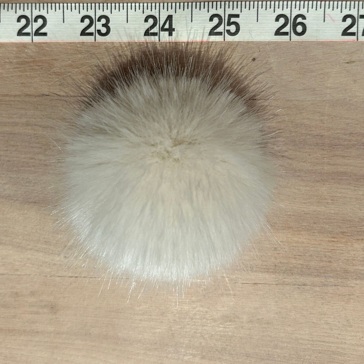 A fluffy pom-pom accessory made of white and light beige faux fur, shown on a wooden surface with a measuring tape at the top for scale.