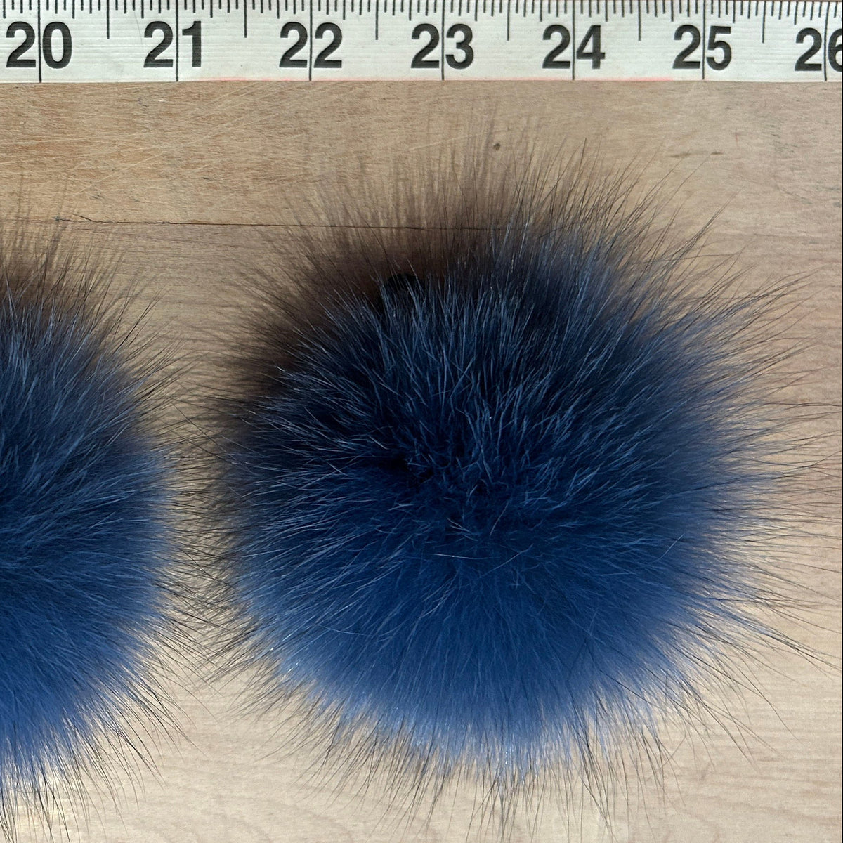 Two fluffy blue fox-fur pom poms placed on a light wood surface with a measuring tape visible at the top for scale.
