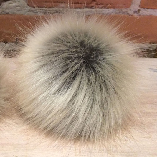 A close-up of a fluffy faux fur pom-pom ball in a light beige and gray colorway, resting on a light wood surface with a brick wall in the background.