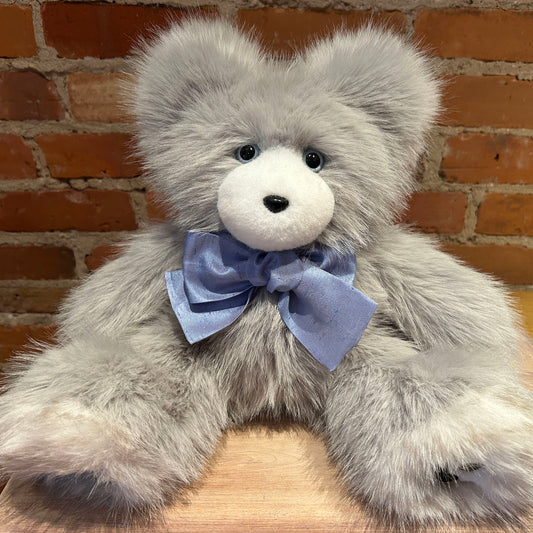 A fluffy light gray teddy bear with a white snout and a light blue satin bow around its neck, posed against a brick wall background.