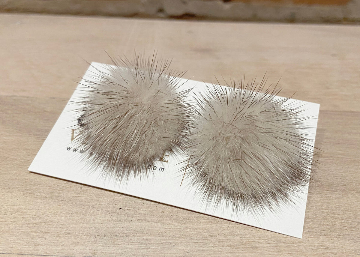 A pair of fluffy pom-pom earrings in a light taupe-gray color, displayed on a white card on a light wood tabletop.