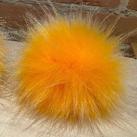 A bright orange fluffy pom-pom made from faux fur or synthetic fiber, shown close up on a light surface with a brick wall in the background.