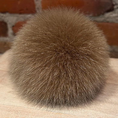 A round, fluffy fox fur pom-pom in a warm brown color, resting on a light wood surface with a blurred brick wall background.