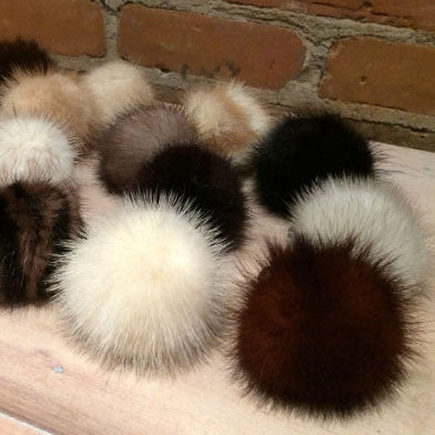 A group of fluffy mink-fur pom-poms in various colors including cream, brown, black, and gray, arranged on a light wooden surface with a brick wall background.