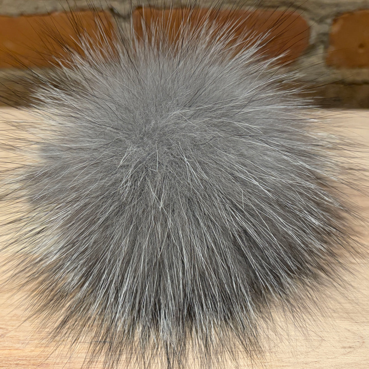 A fluffy gray silver fox fur pom-pom purse pom accessory resting on a light wood surface with a blurred brick wall background.