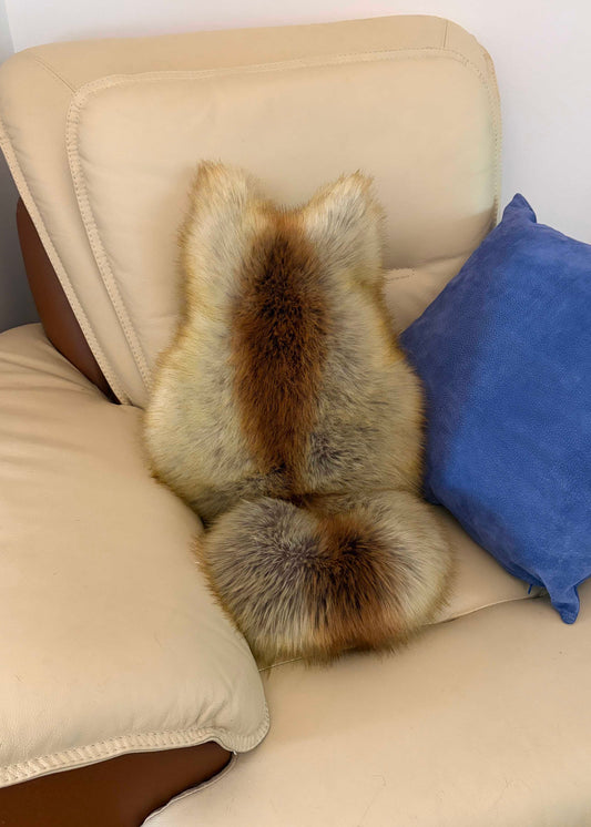 A fluffy faux-fur or animal-pelt style cushion shaped like a fox or dog, shown from the back, resting on a cream leather chair next to a blue pillow.