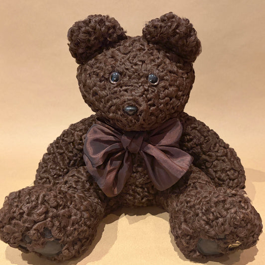 A brown persian lamb teddy bear with shiny dark eyes, a black nose, and a dark purple satin bow around its neck, sitting against a plain beige background.