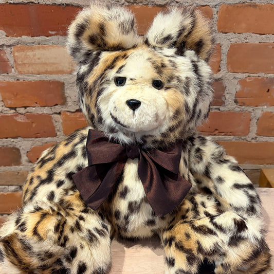 A plush teddy bear with a spotted cheetah-like pattern in cream, tan, and black, wearing a dark brown bow, posed against a red brick wall background.