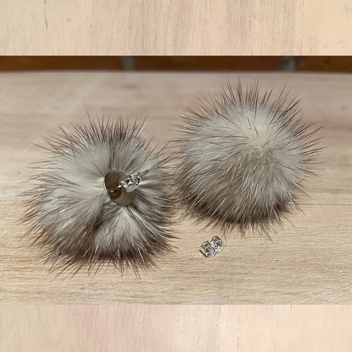 A pair of fluffy pom-pom stud earrings made from light gray and brown mink  fur, with small silver-toned metal posts and clear rhinestone studs, displayed on a light wood surface.
