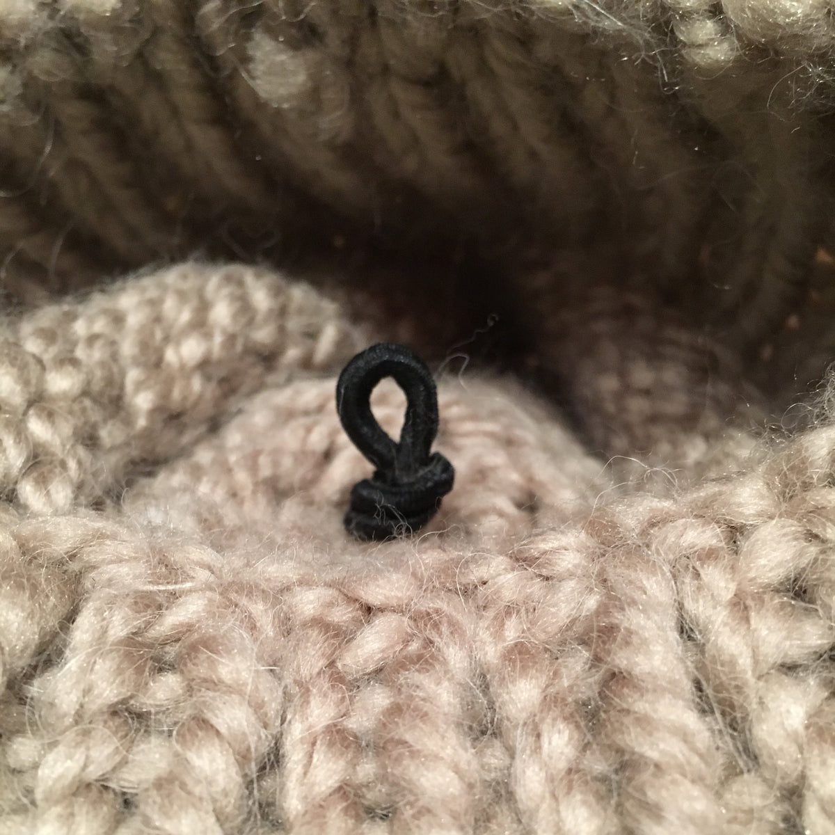 Close-up of a chunky knit textile in a warm taupe-beige color, showing the texture of the yarn and a small black loop detail at the center.