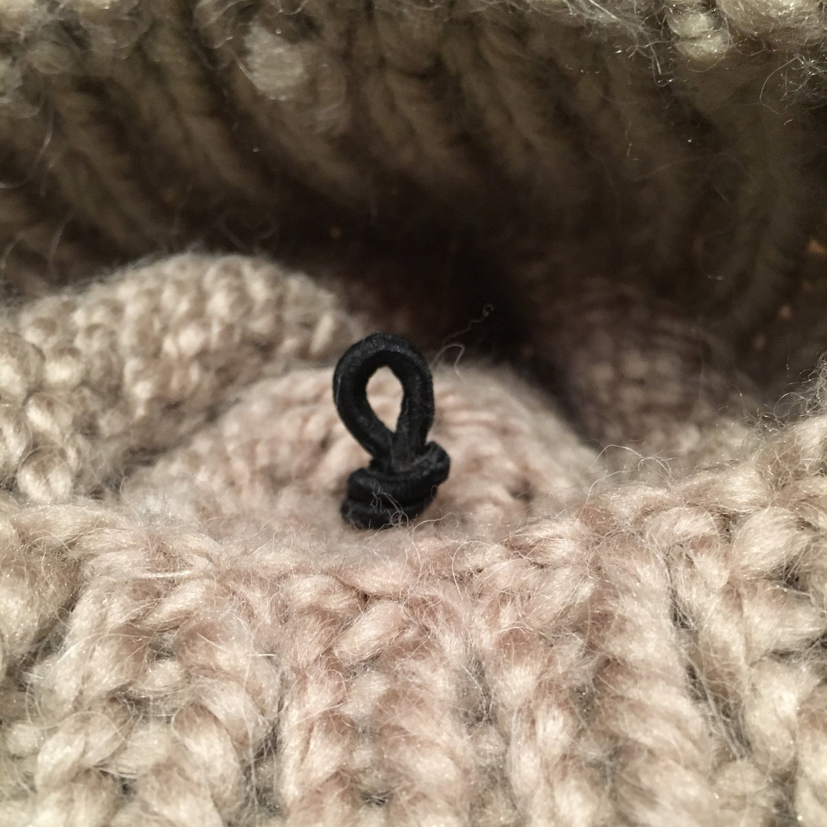 Close-up of a chunky knit textile in a warm taupe-beige color, showing the texture of the yarn and a small black loop detail at the center.