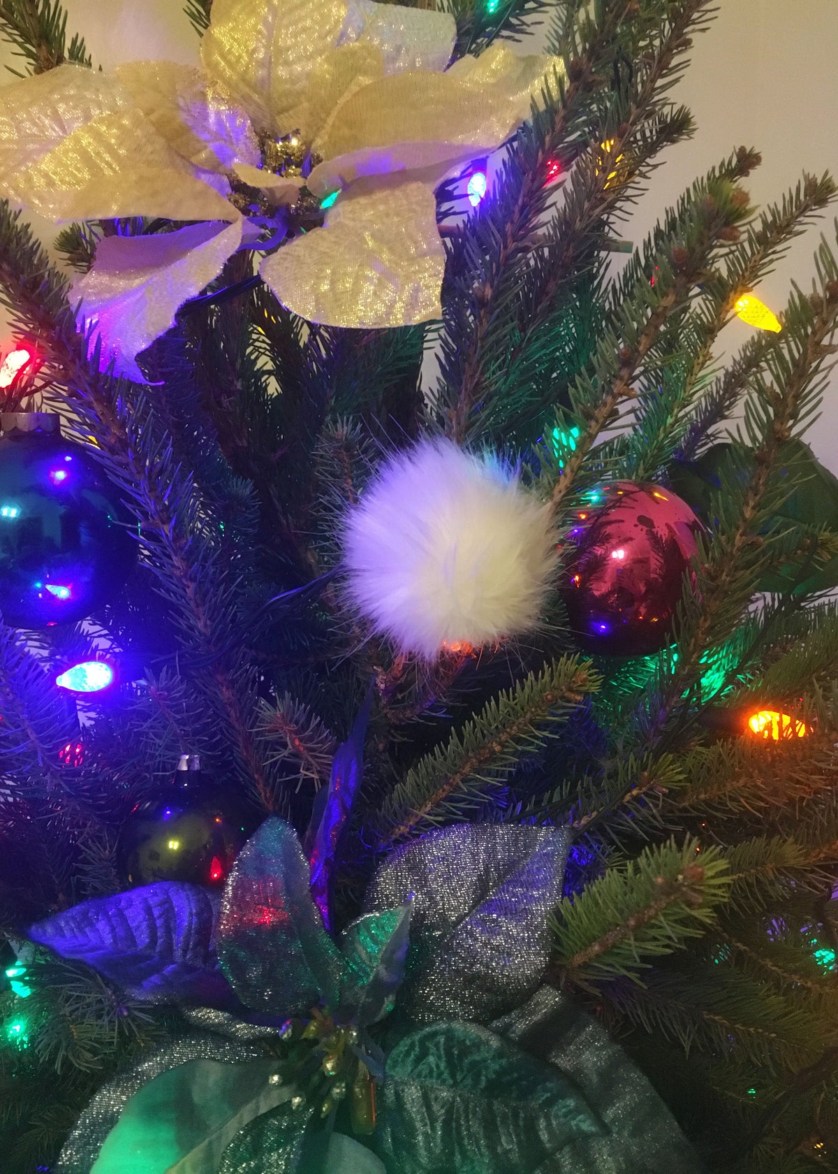 Close-up of a decorated Christmas tree with green pine branches, multicolored string lights, a white fluffy ornament, shiny red and blue baubles, and a large gold poinsettia topper.