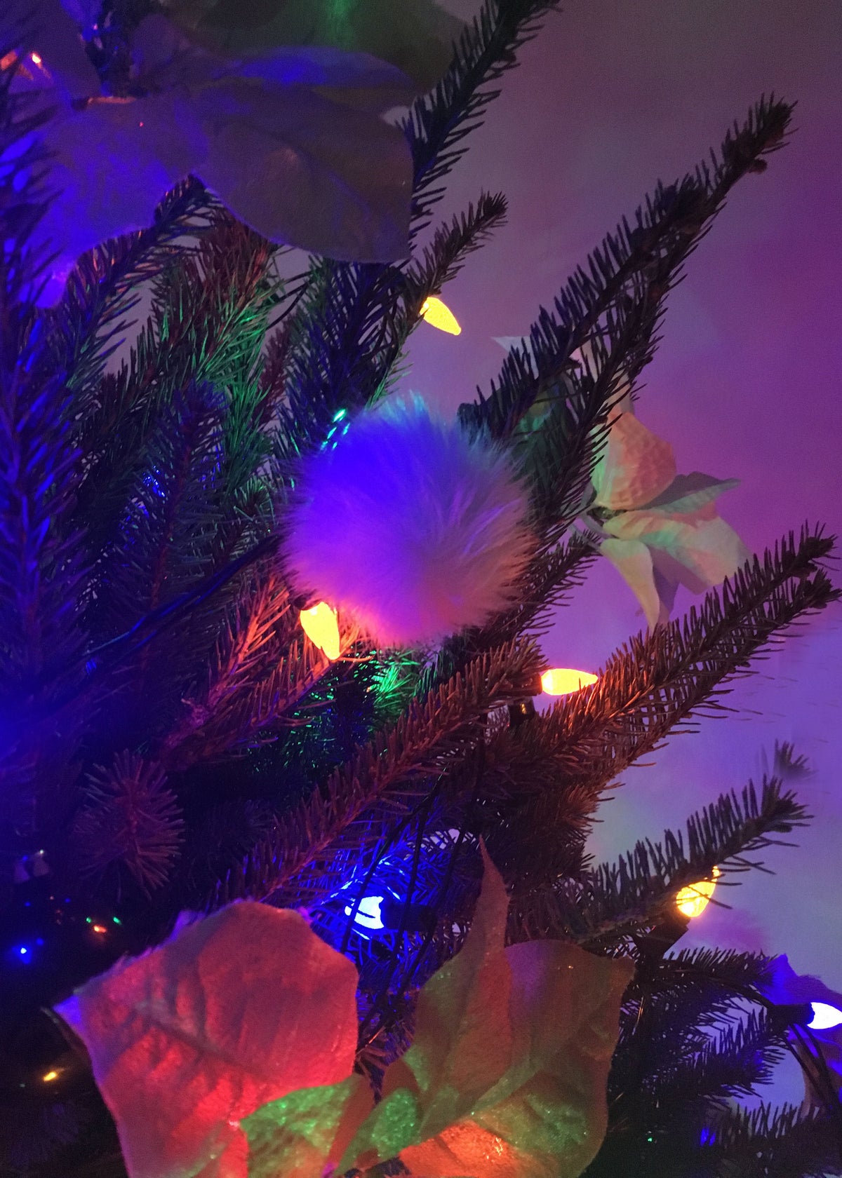 A close-up of a Christmas tree decorated with multicolor fairy lights, a fluffy white pom-pom ornament, and large paper leaves under colored ambient lighting.