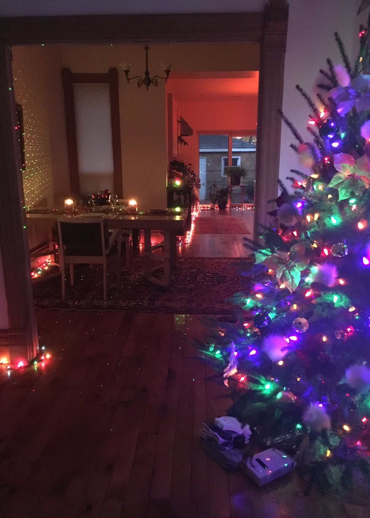 A decorated Christmas tree with multicolored lights stands in a dimly lit living room, with a dining room and fireplace visible in the background.