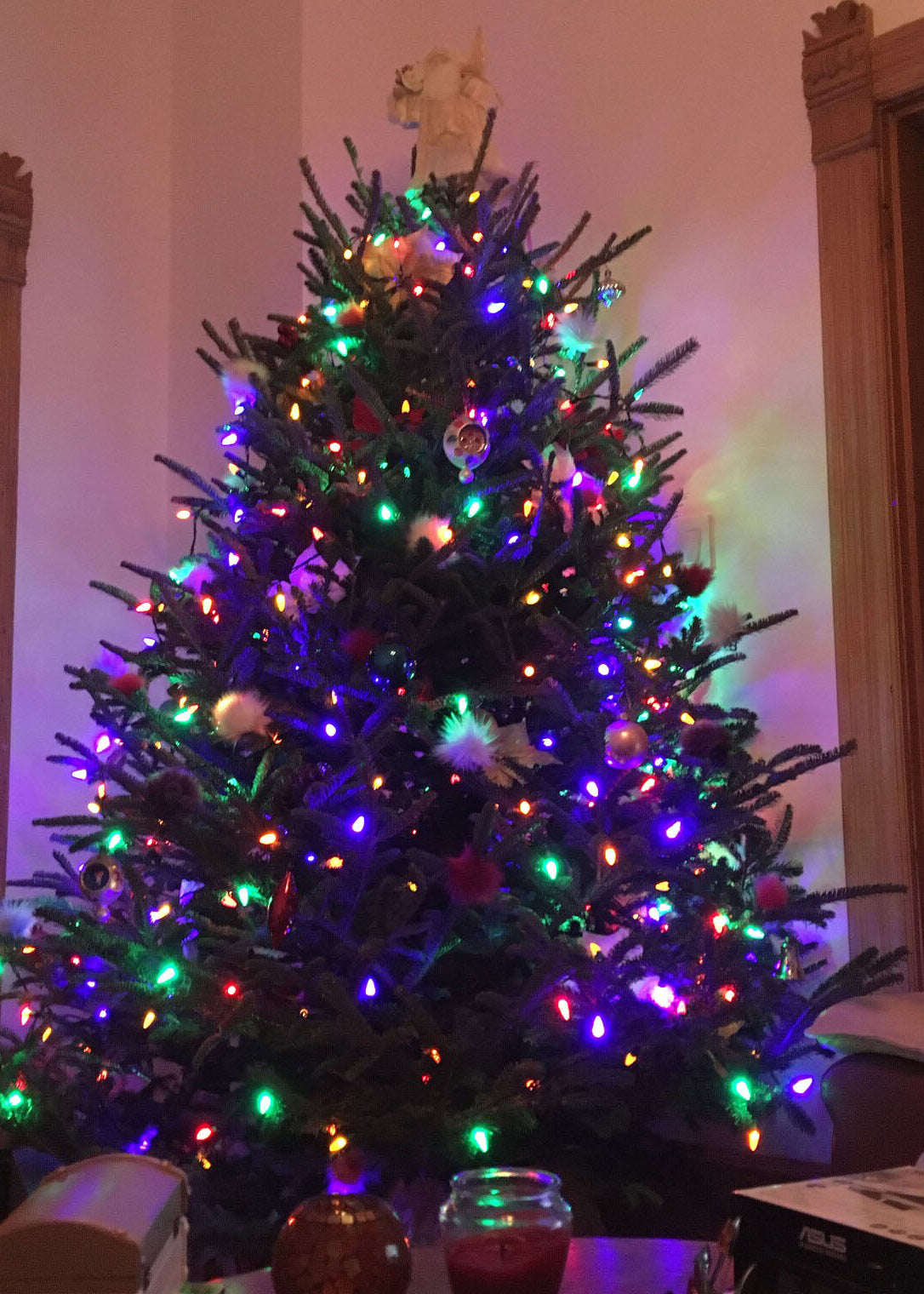 A decorated Christmas tree with multicolored string lights and ornaments, standing indoors against a wall with wooden door frames visible on both sides.