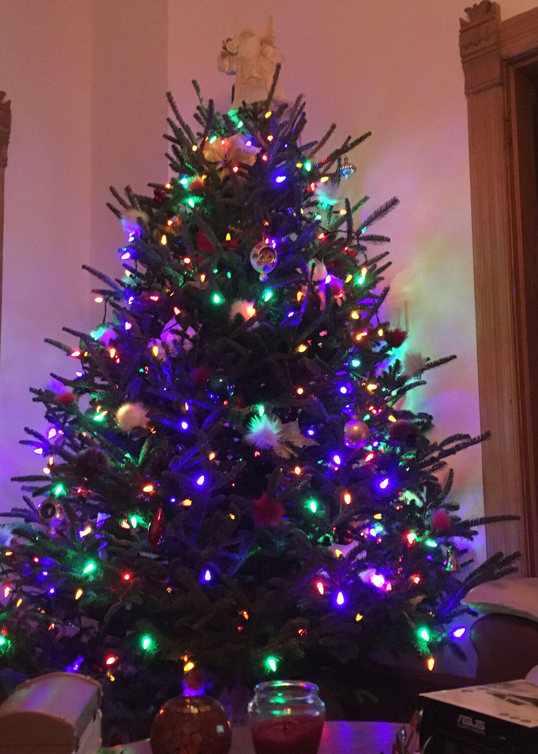 A decorated Christmas tree with multicolored string lights and ornaments, standing indoors against a wall with wooden door frames visible on both sides.
