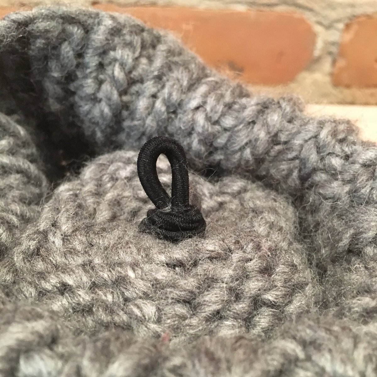 Close-up of a gray knit or crocheted textile item with a small black loop fastener attached, set against a blurred brick background.