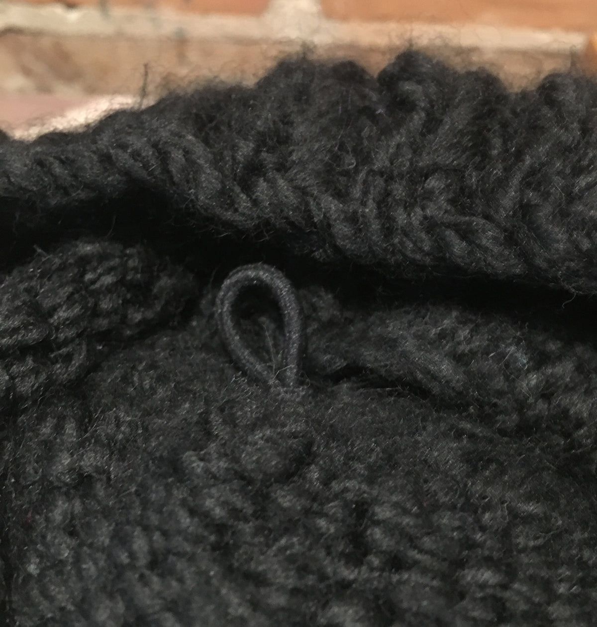 Close-up of a chunky black knit fabric with a small loop detail, set against a blurred brick background.
