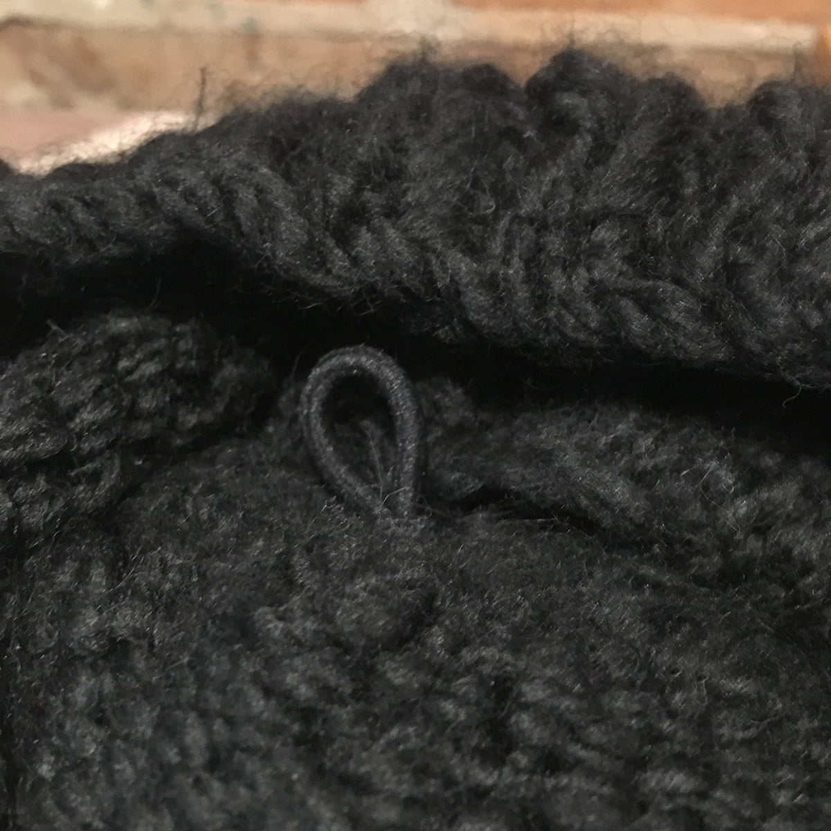 Close-up of a chunky black knit fabric with a small loop detail, set against a blurred brick background.