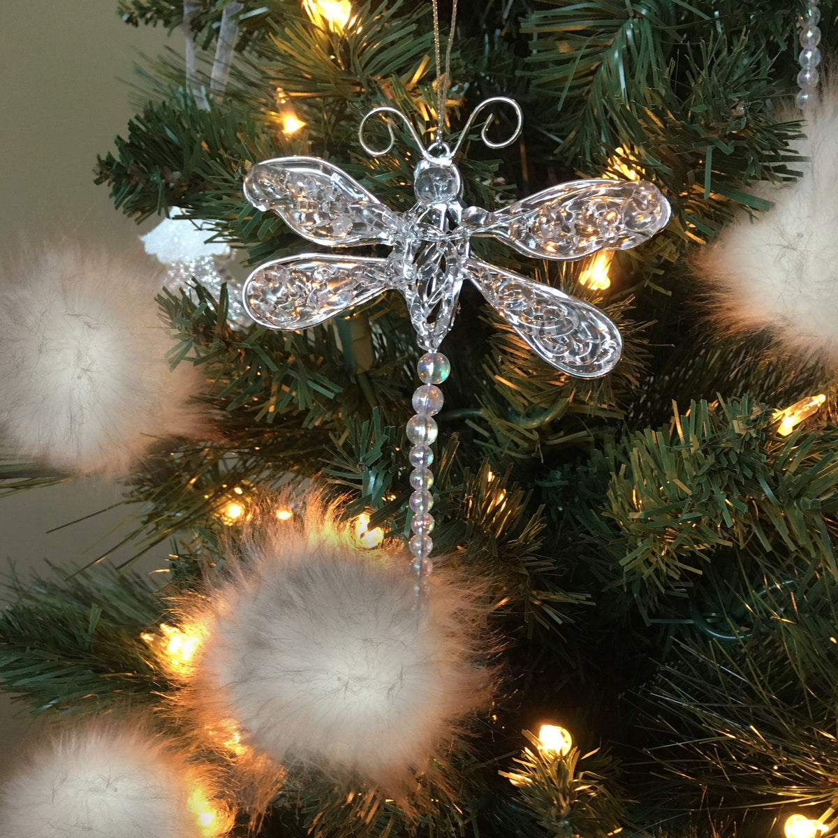 A clear, ornate dragonfly Christmas ornament with intricate cutout detailing and a hanging beaded tail, displayed on a green Christmas tree with warm white lights and fluffy white pom-poms.