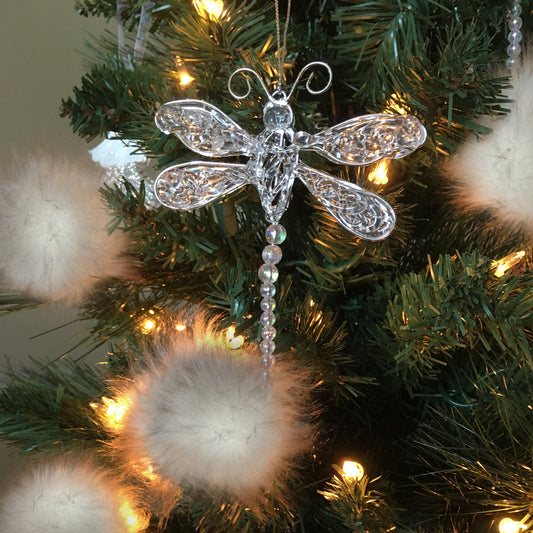 A clear, ornate dragonfly Christmas ornament with intricate cutout detailing and a hanging beaded tail, displayed on a green Christmas tree with warm white lights and fluffy white pom-poms.