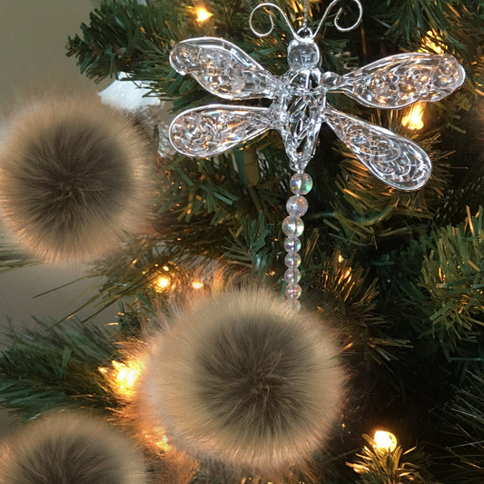 A clear, textured dragonfly Christmas ornament hanging on a green Christmas tree with warm white lights and fluffy beige pom-poms.
