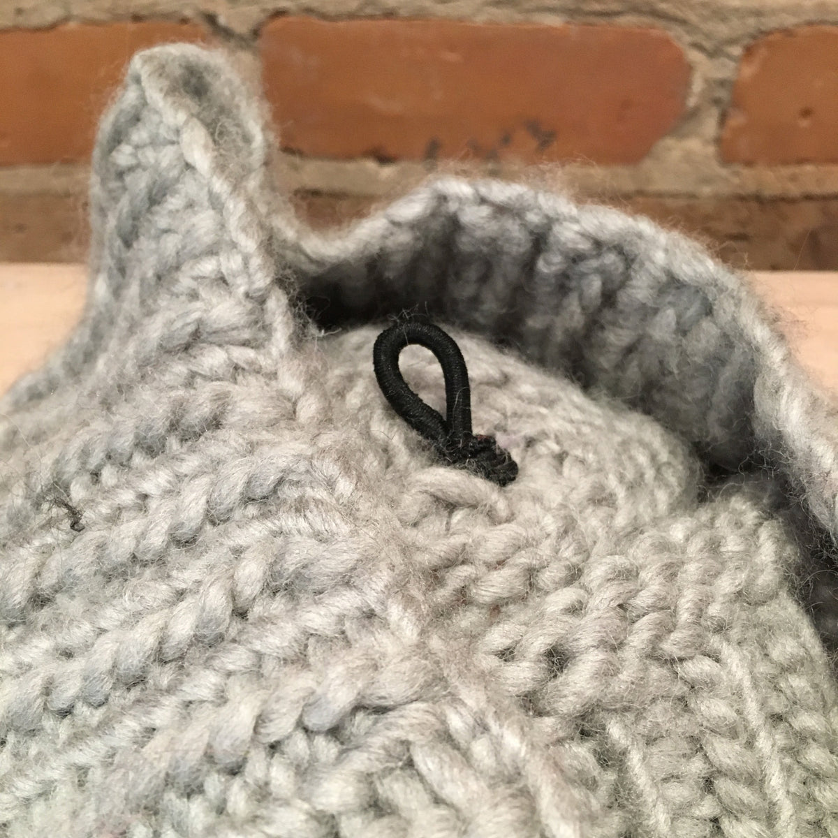 Close-up of a chunky knit light gray garment, showing the textured stitch pattern and a small black loop detail, with a brick wall in the background.