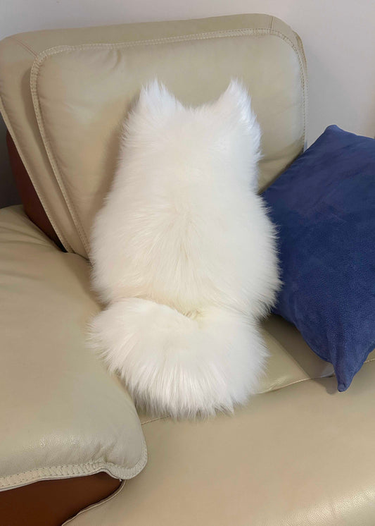 A fluffy white faux fur cushion shaped like a cat, shown from the back resting on a beige leather armchair next to a blue pillow.