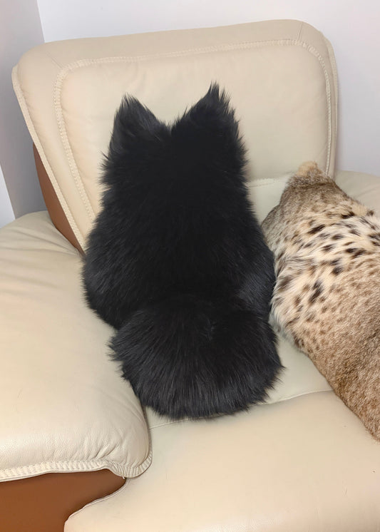 A fluffy black cat shaped pillow sits on a cream-colored leather armchair, viewed from behind. A leopard-print cushion rests on the right side of the chair.