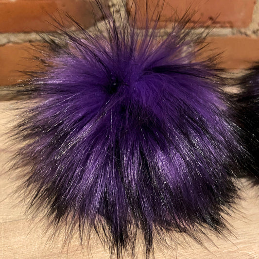 A close-up of a fluffy faux fur pom-pom accessory in vibrant purple with black tips, resting on a light wood surface against a brick wall background.